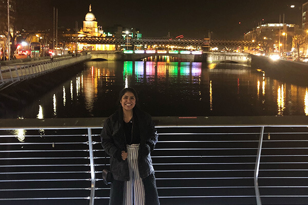 A student stands in front of a bridge in Dublin, lit with city lights at night